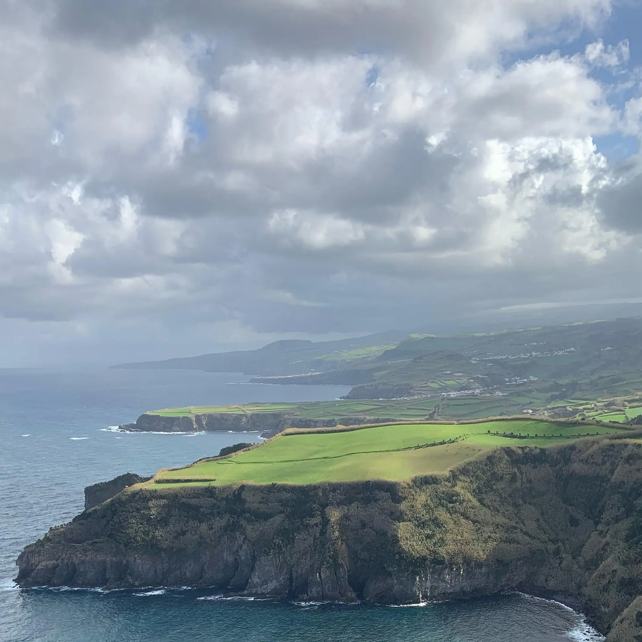 Green fields atop coastal cliffs along the Atlantic Ocean in Portugal, with rolling hills in the distance.